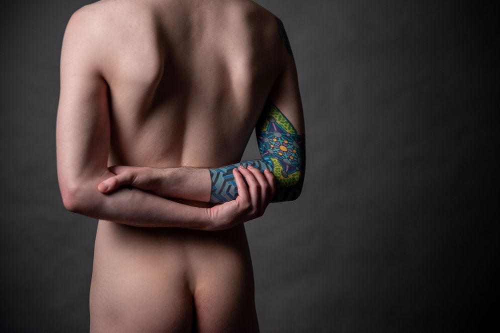 From behind we reveal he is nude, his butt in frame and arms crossed behind his back - clutching his tattoo’d forearm