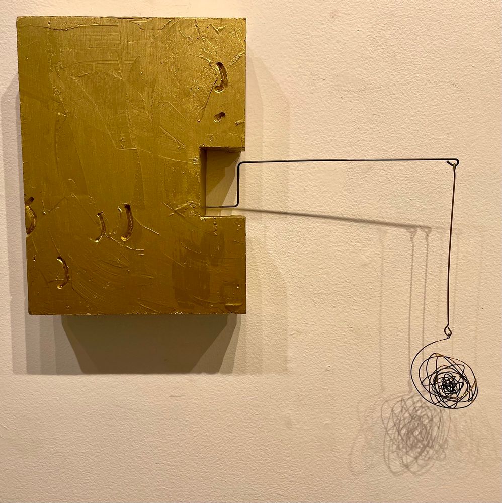 Gold painted box form hanging on wall with steel wire extension coming out of box to the right. Brass wire dangling down from steel wire at right angle, black wire scribble form hanging from brass. The sculpture casts dynamic, layered shadows on wall.