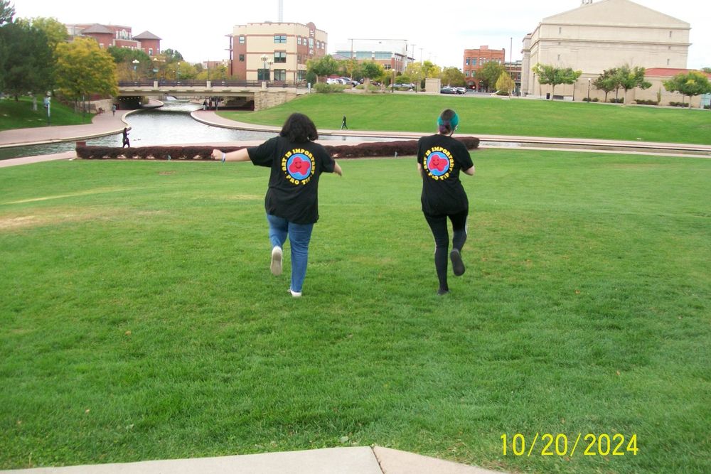 A photo of Everest (left) and Grace (right) skipping in a green grass field with their backs facing the camera. There is a river to the left and in front of them. They are both wearing black, Dan and Phil “Don’t Cry, Craft” shirts, with the text “Art is important pro tip” curved around a blue circle, encompassing a red blob with a smiley face. Everest has shoulder length brown hair, and they are wearing blue jeans and white shoes. Grace is wearing their brown, teal, and purple hair in a low bun, and is wearing black leggings and black crocs. There is a date stamp on the bottom left reading 10/20/2024.