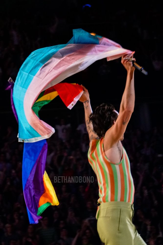 harry styles live on stage. waving a trans flag and a rainbow flag 