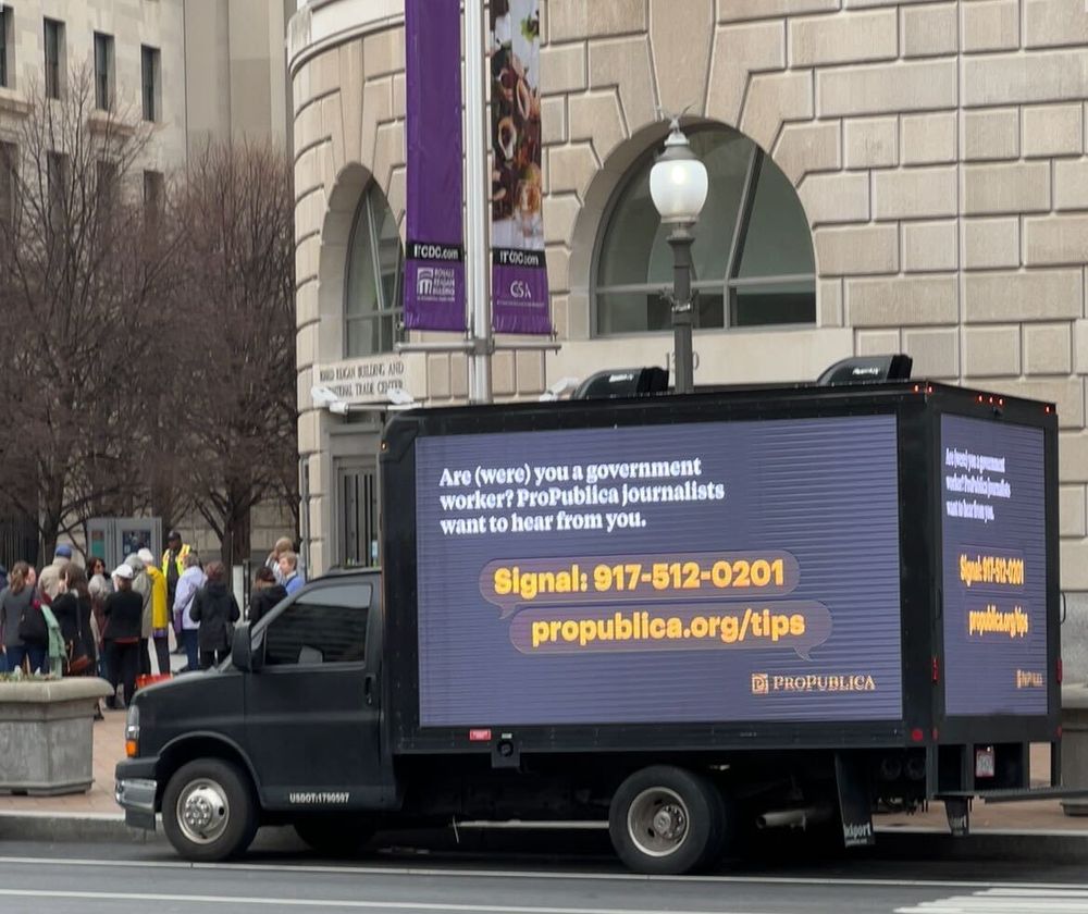 A black mobile billboard truck parked near the Ronald Reagan Building and International Trade Center in D.C. On the side and back of the truck, a LED screen reads: Are (were) you a government worker? ProPublica journalists want to hear from you. Signal: 917-512-0201. propublica.org/tips.