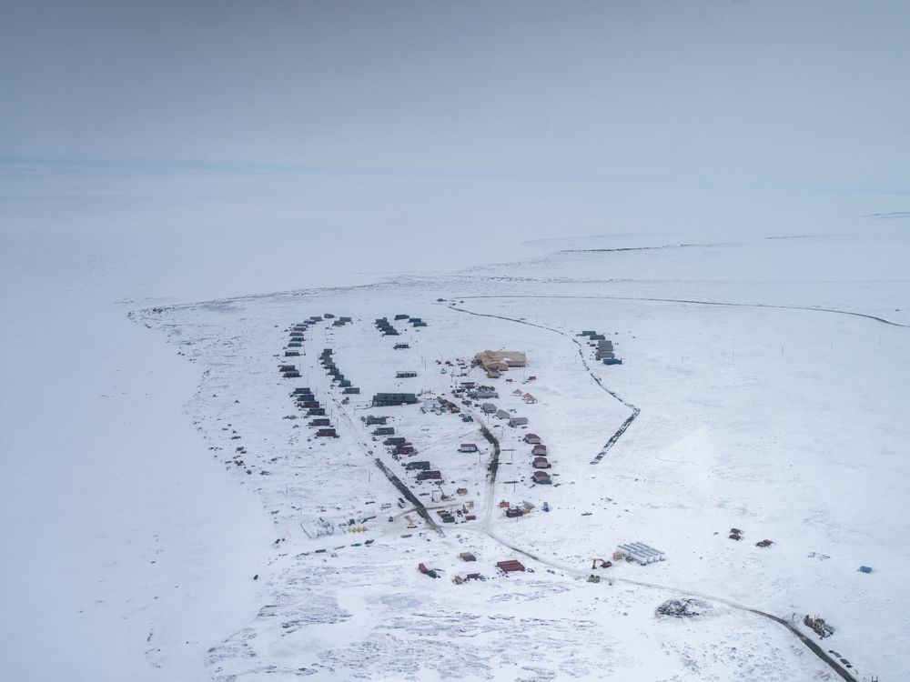An aerial view of Metarvik, Alaska, a small village of several dozen buildings in the middle of a vast snowy expanse.