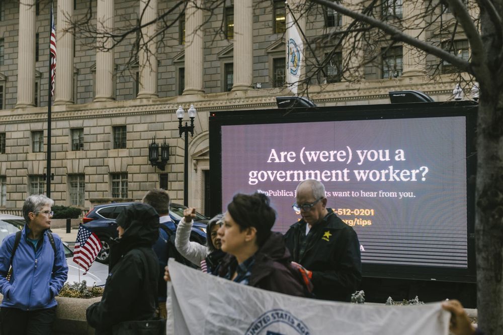 Multiple demonstrators stand near USAID’s former headquarters at the Ronald Reagan Building and International Trade Center, some holding a giant white flag with the USAID logo. Behind them is a a parked mobile billboard truck. Text on the side of the truck begins with: "Are (were) you a government worker?" The rest of the text is partially covered by the demonstrators, but shows contact information for ProPublica.