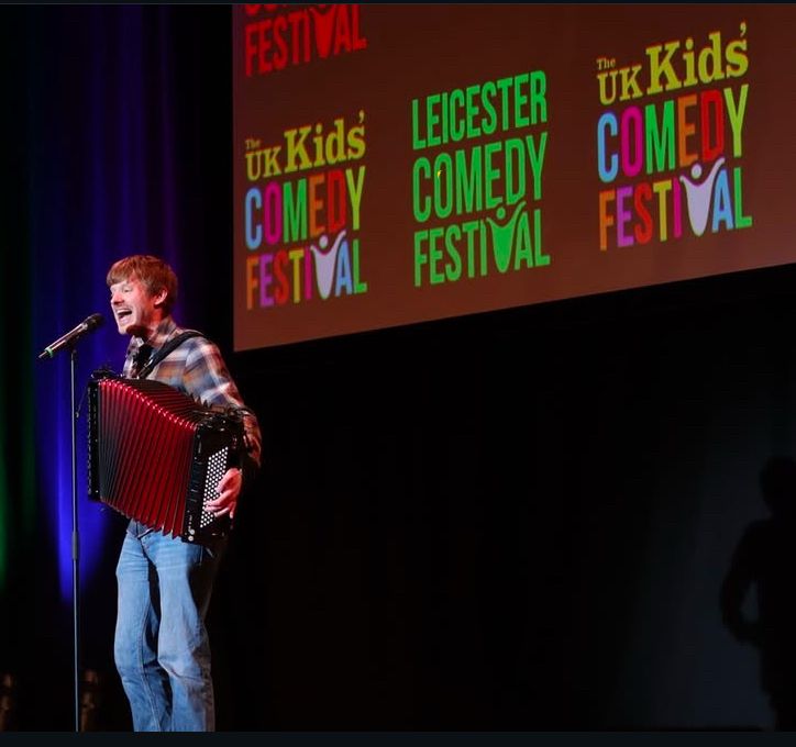 A picture of David with his accordion in full flow at the mic in front of a big screen with the Leicester Comedy Festival logo and UK Kids Comedy festival logo 