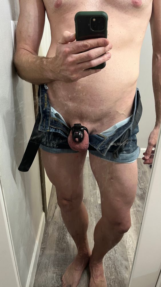 Nude caucasian cisgender male from lower chest down to feet standing in front of a mirror with phone-camera visible at chest level. Wearing unzipped/opened jean shorts that have the fly spread to reveal penis in kink3d chastity cage.