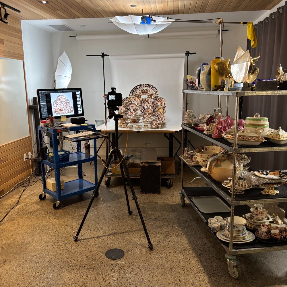 Part of a photographer's studio set up for tabletop product photography. The table is covered with a white paper backdrop with a 66-piece Royal Crown Derby "Imari" dinner service set up on it. There is also a rolling cart nearby with a whole bunch of other decorative ceramic and glass things on it. I'd love to smash all of it.