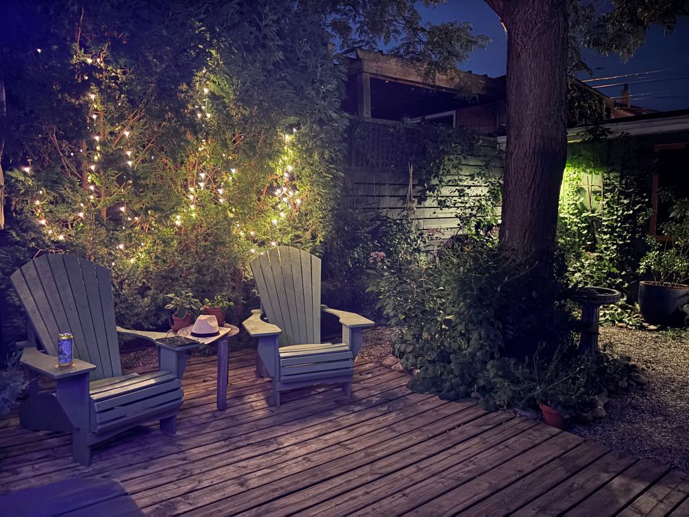 Another angle showing some cedars filled with faerie lights and a couple of Muskoka chairs on a wooden deck.