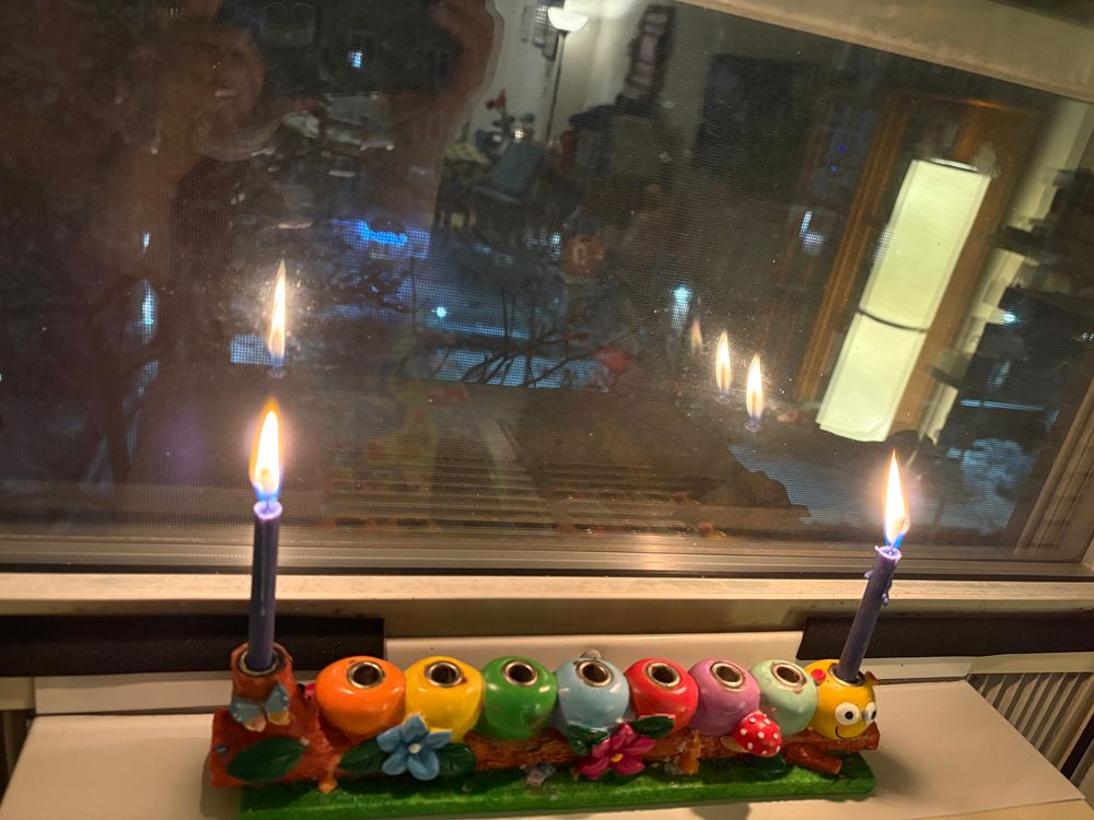Brightly colored menorah in the shape of a caterpillar sitting on a log with flowers, butterflies, and mushrooms. 