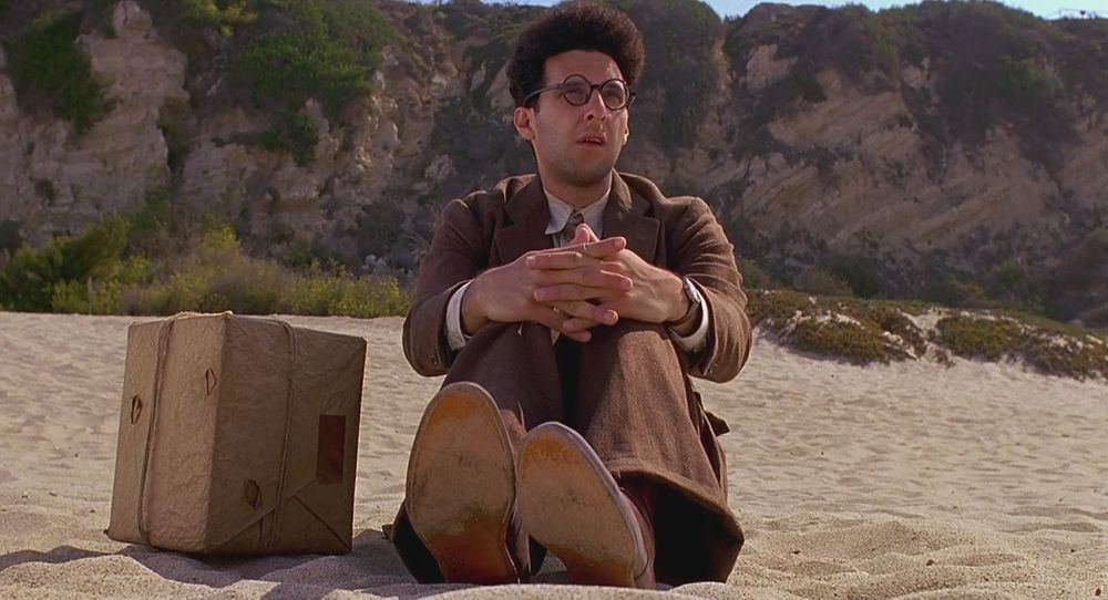 John Turturro (wearing a suit) sitting on a beach with a box. 
