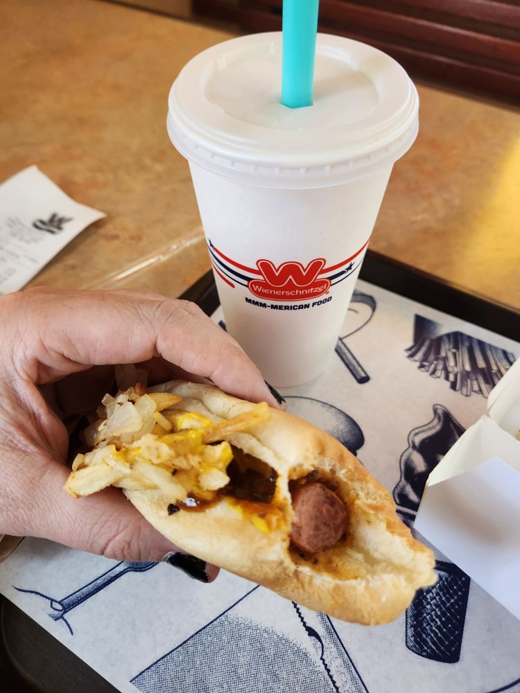 Ricki holding a fast food hot dog with chili cheese fries on it next to a shake.