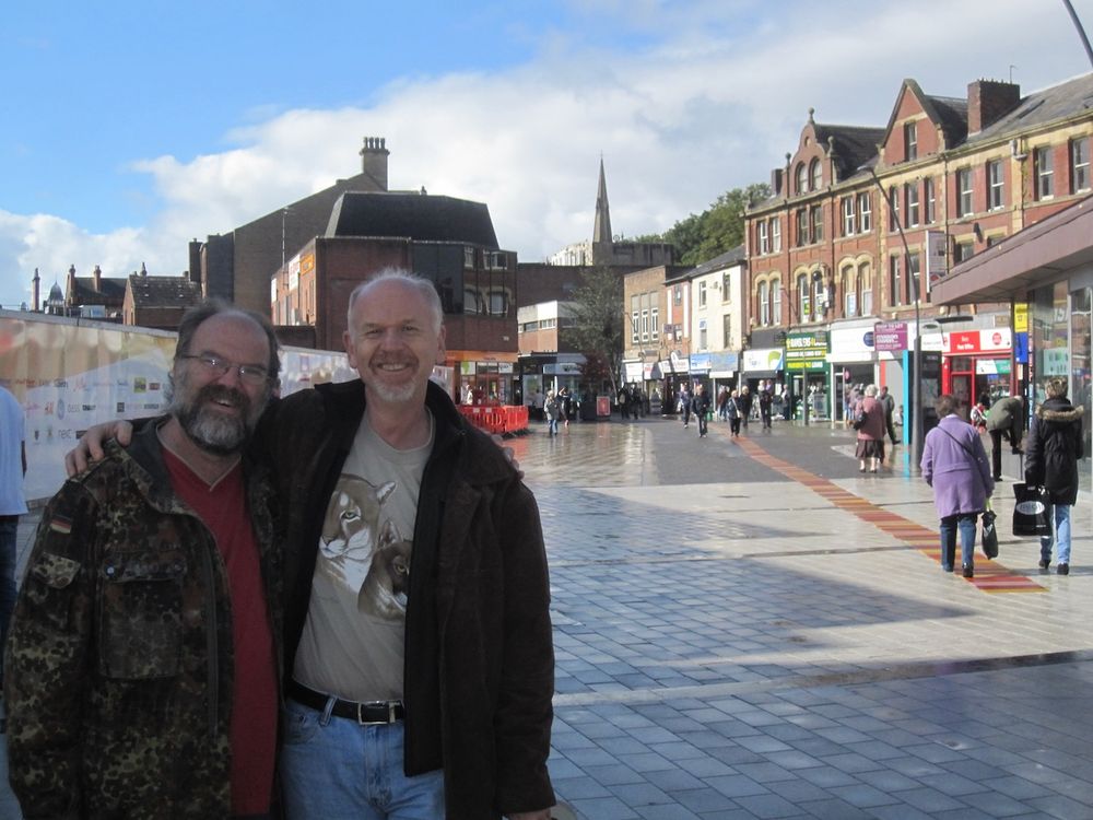 Two men stand in front of a plaza where people are wandering to shops and market stalls. They're grinning broadly. It's a beautiful day.
