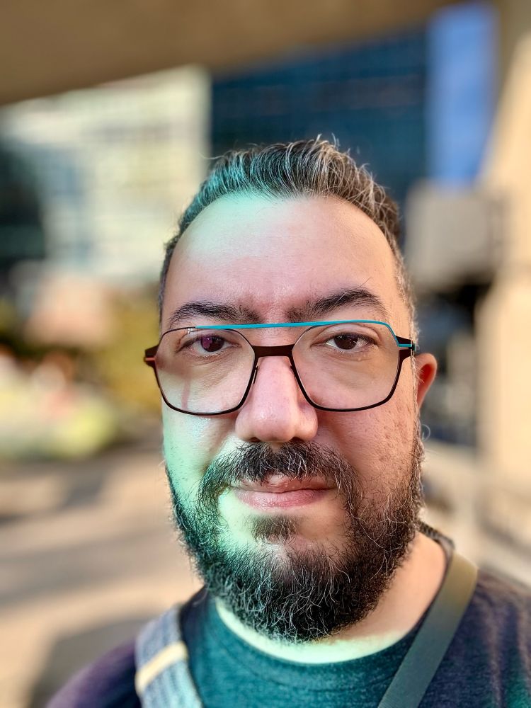 A photograph of a bearded, bespectacled face with a neutral expression against a very blurry background.