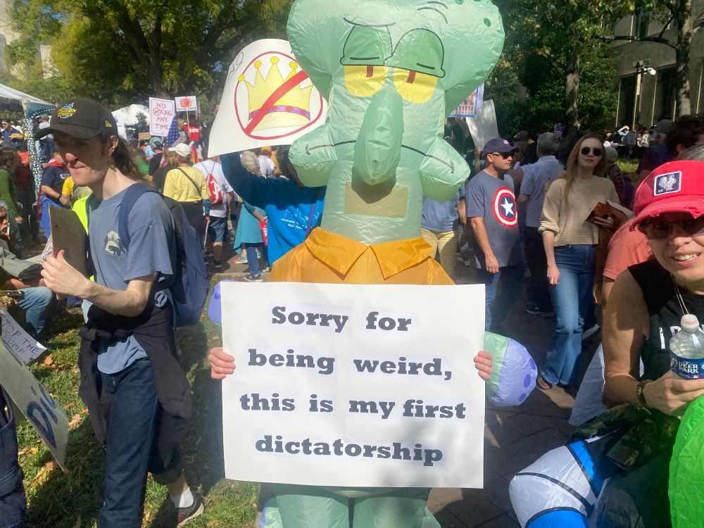 A protester in a Squidward inflatable costume holds a white sign with black letters at the #NoKings rally. It says, "Sorry for being weird, this is my first dictatorship..." #NoKings