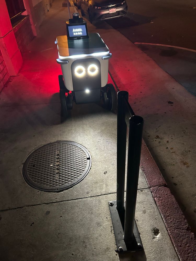 A food delivery robot confused by an empty sidewalk bike rack, unclear how to move forward, no programming on how to go back. Who among us. 