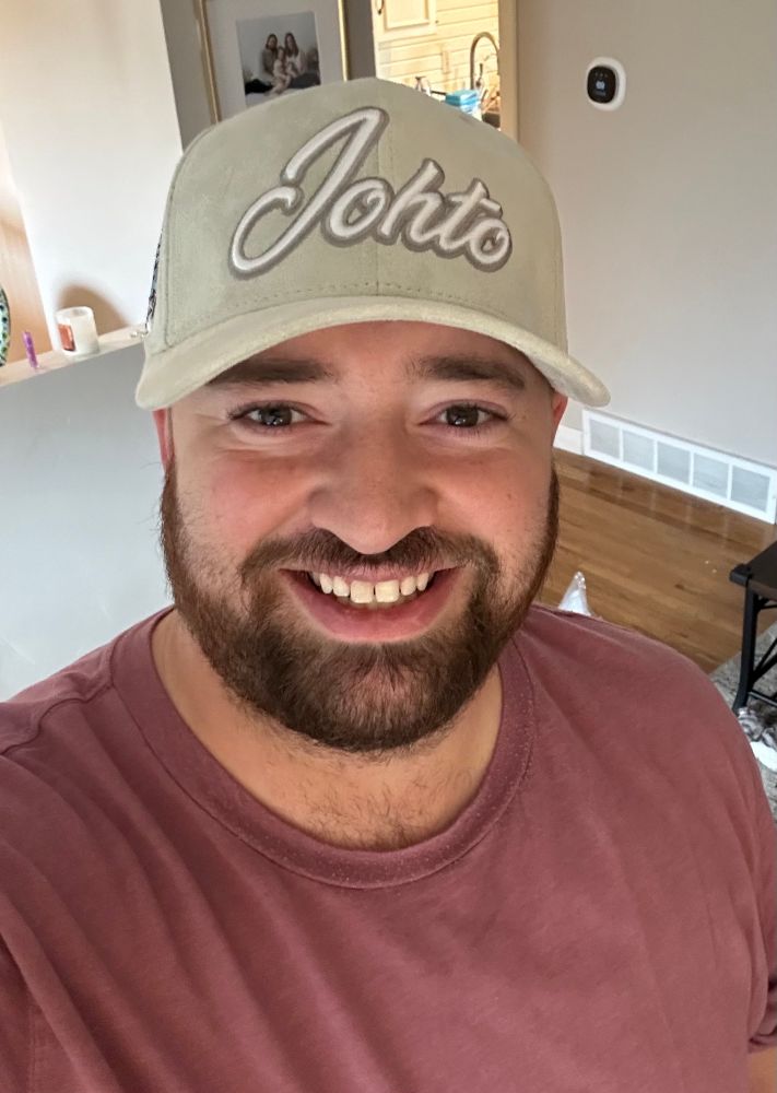 Picture of myself smiling while wearing a dark pink shirt and tan hat with the word Johto on the front.