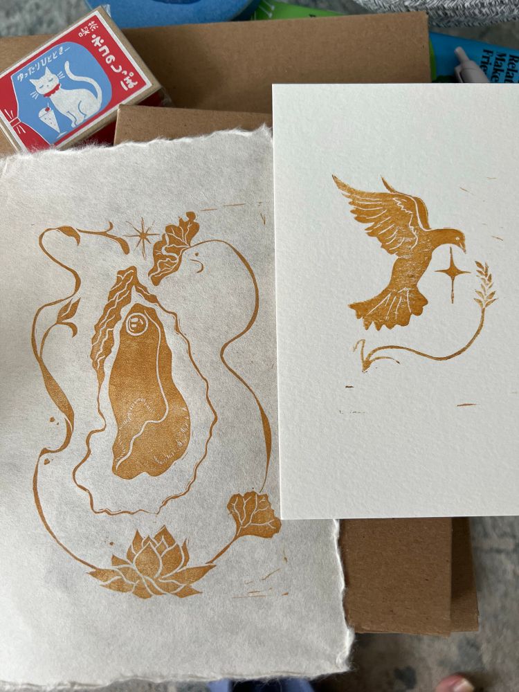 A photo of two linocut prints. One is a copper oyster with lotus blossoms and the other is a copper dove with a sheaf of wheat.