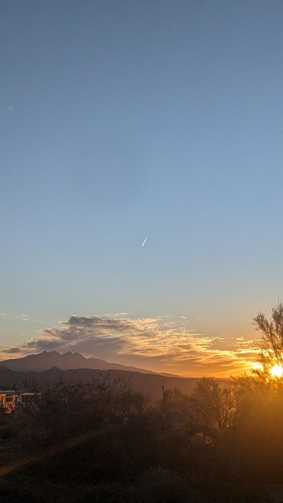 A desert sunset scene: the sun glows low at the right edge, coloring wispy clouds orange above layered mountain silhouettes. Dark cacti and scrub fill the foreground