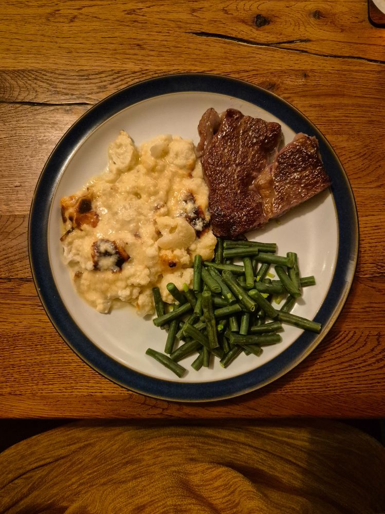 Japanese wagyu steak with cauliflower cheese, and green beans. On white plate with blue rim, on wooden table.