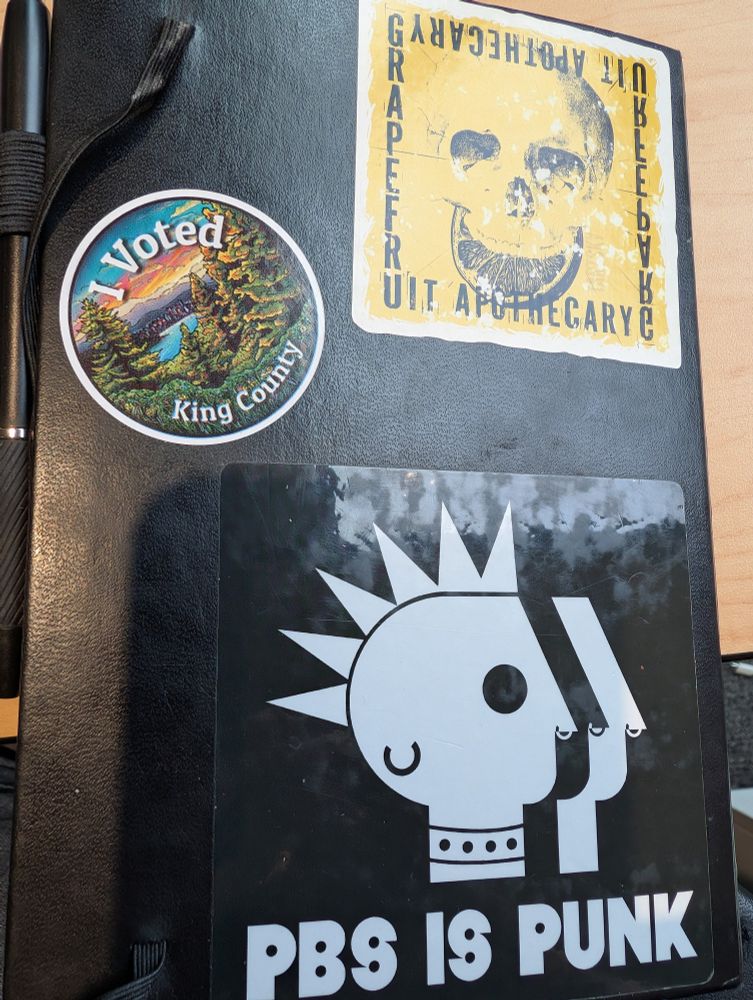 Black notebook with three stickers, one says I voted King County, one says PBS is punk, and one's a skull saying Grapefruit Apocalypse