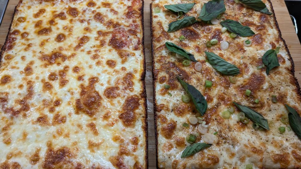 Two Detroit style pizzas; plain cheese on the left, a pizza with Thai basil, sliced green onions, and a mala seasoning blend sprinkled on top on the right.