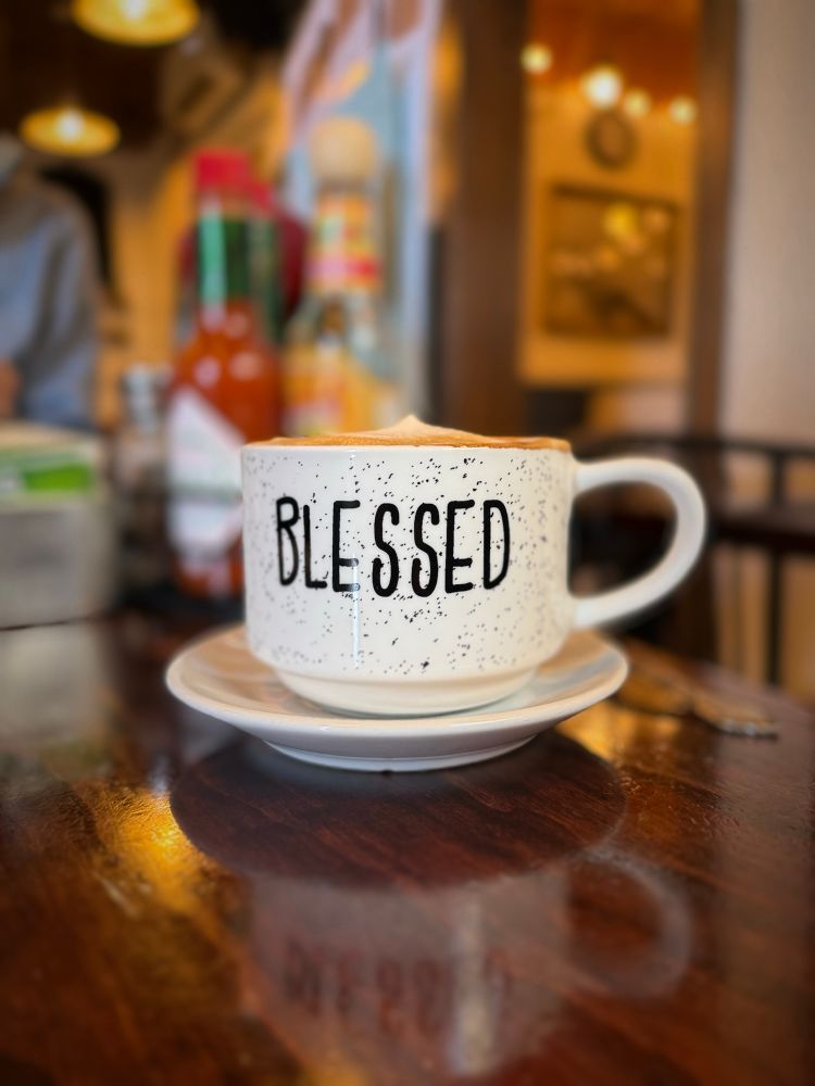 A white mug filled with a cappuccino. It sits on a brown wooden table. The word “Blessed” is printed on the mug in black letters. 