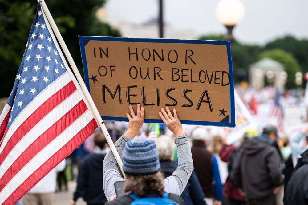 Person at the MN No Kings protest holding a sign that says "In honor of our beloved Melissa" followed by another person holding an American flag.