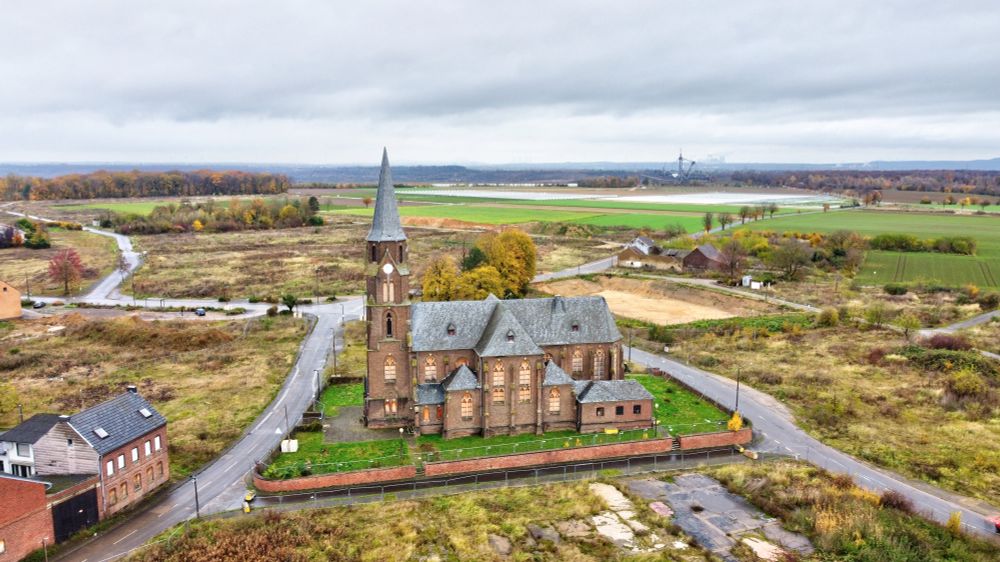 Luftbild der Manheimer Kirche in Großaufnahme. Äußerlich intakt, zeigen sich Verfallserscheinungen. Die Fenster wurden durch Bretter ersetzt, die Glocken wurden herausgerissen, die Uhren sind zu unterschiedlichen Zeiten stehengeblieben. Die Kirchenmauer steht, alle Bäume sind gefällt. Die meisten Häuser wurden dem Erdboden gleichgemacht. Links vorne sind noch zwei zu sehen. Im Hintergrund der Sündenwald (ca. 1km) und ein Braunkohlebagger (knapp 2km). Grüne Felder, weiße Abdeckungen von Erdbeerfeldern.