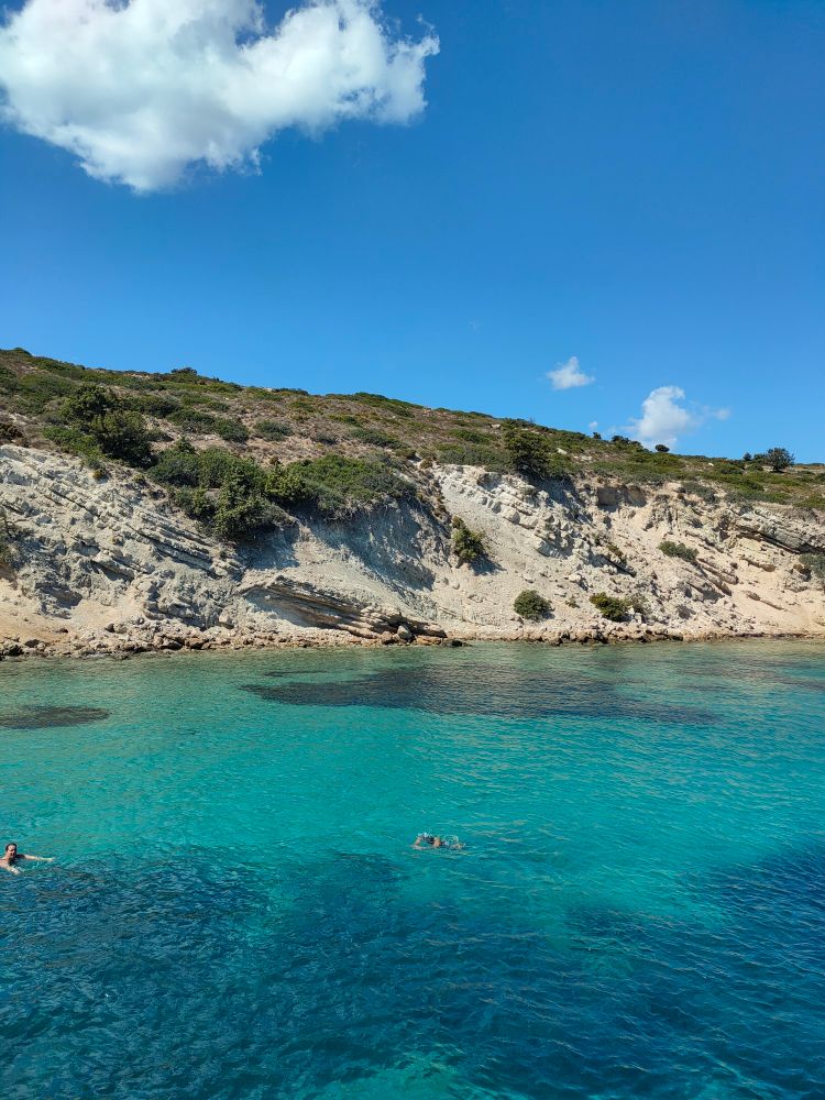 Mediterranean cliff shoreline with a blue sky and clear blue green water with a few swimmers.