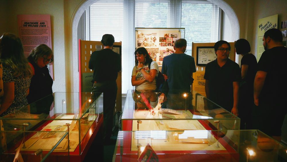 The audience peruse the exhibition, which has four red cases in the centre of the room and panels around the outside.