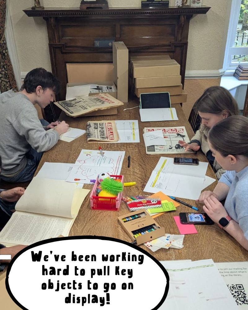 A group of volunteers sat around a table in the WCML reading room, looking at Big Flame material. The caption reads "We’ve been working hard to pull key objects to go on display!"