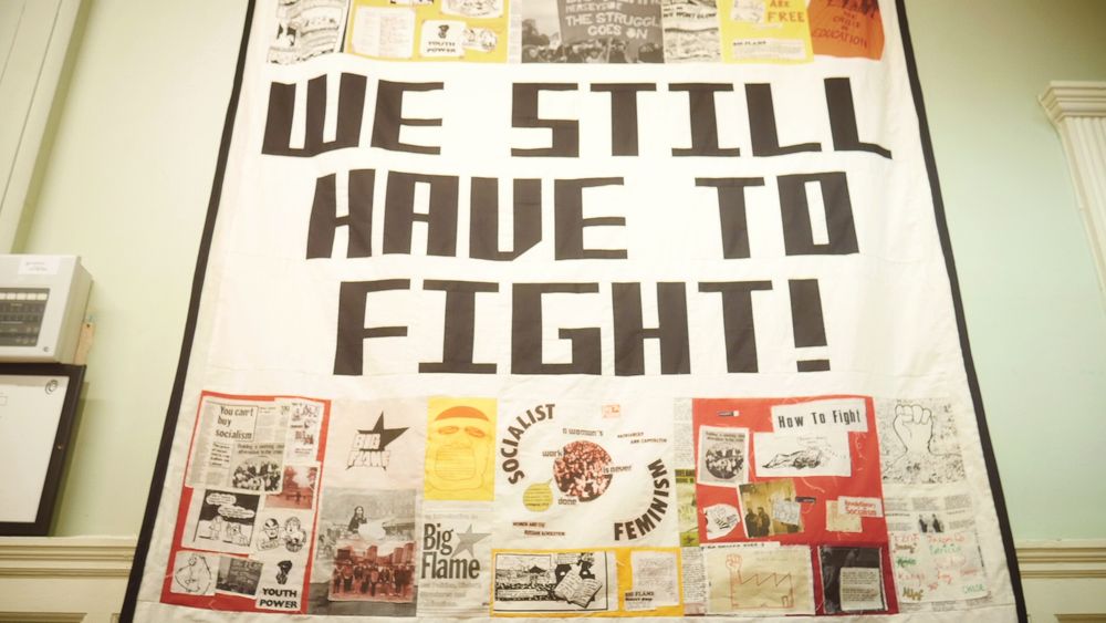 A close-up of the Little Flames banner, which reads ‘we still have to fight!’ in the centre and has collaged fabric images around it.