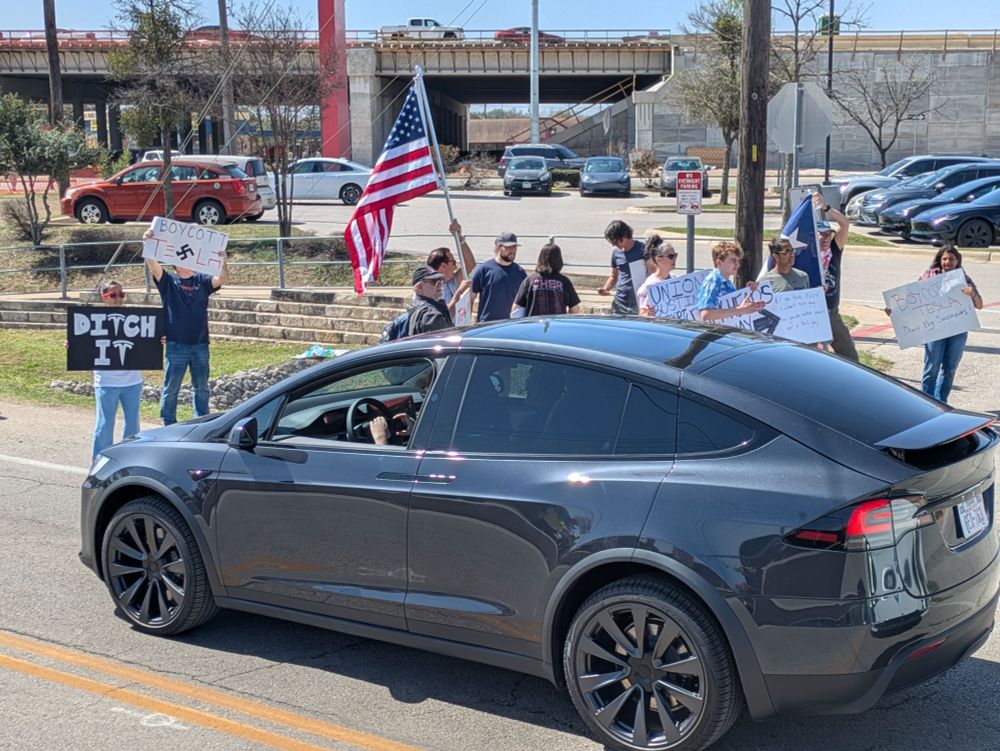 A woman with a sign that says "Ditch It" stands next to a Tesla car sitting in traffic