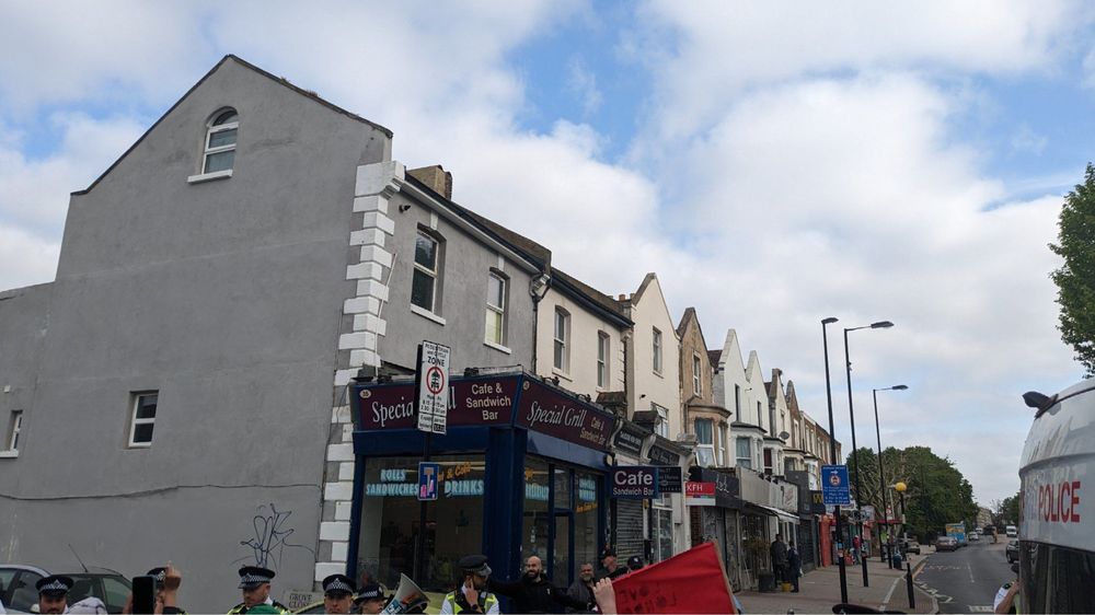 A photo of a small shop called special grill cafe and sandwich bar with three  bald men stood in front and surrounded by police. In front of them is a red banner and megaphone facing them.