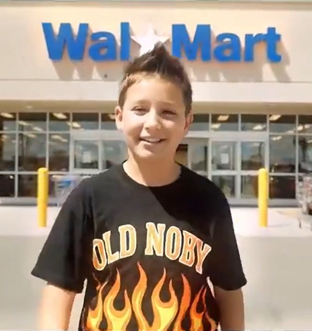 Uncanny AI-generated child standing in front of a Wal-Mart and wearing a black t shirt with sick flames and Old Noby written on it.