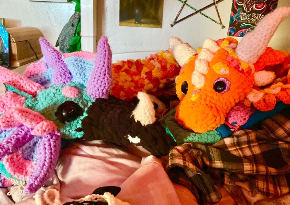 A pair of crocheted dragon heads look at the camera. The one on the left is blue, pink, purple, and black with fins and a beak. The one on the right is orange and yellow with multicolored scales. 