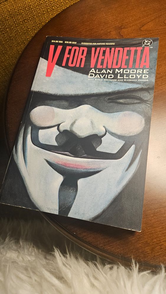 The graphic novel of "V for Vendetta", by Alan Moore and David Lloyd, featuring a painting of the grinning "V" mask on the cover, sits on a round, wood table.