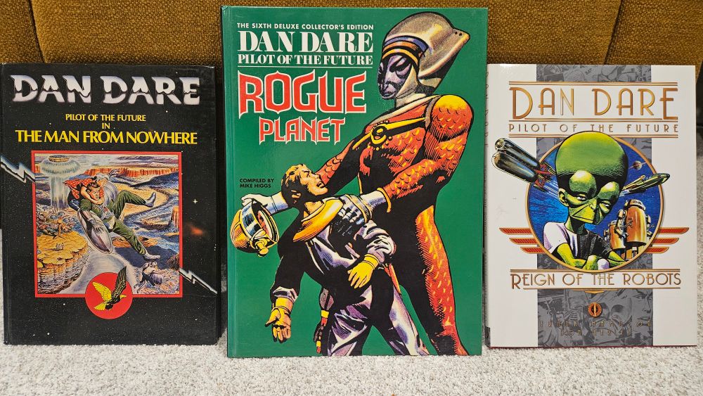 3 reprint volumes of Dan Dare comic strip stories: a small, black hardcover with "futuristic" title fonts and a single panel of Dan Dare and Digby floating down to an alien world; a large green hardcover in the centre with Dan being apprehended by a giant, stern-faced alien; a white hardcover with retro art deco design, featuring a picture of the green-skinned Mekon in a circular frame.