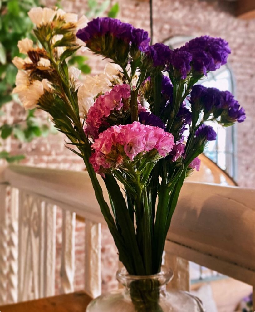 Small bouquet of white, purple and pink flowers in a tiny vase, on a table adjacent to the (inside) wooden balcony railing that looks out on a mirror filled brick wall