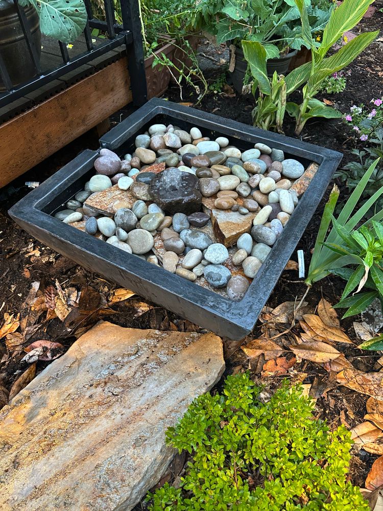 Square pond water feature filled with rocks with a single water fountain coming out of a rock in the middle. No standing water is visible. 