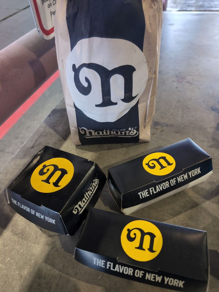 Three Nathan's boxes sit on a stone bench in front of a brown paper bag with Nathan's branding. The boxes have the Nathan's logo and say "the flavor of New York."