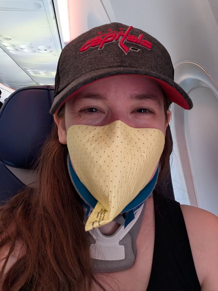 Emily is on an airplane smiling beneath her mask. She is wearing a yellow stick-on N95 mask, a blue and white neck brace, and a Washington Capitals hat. 