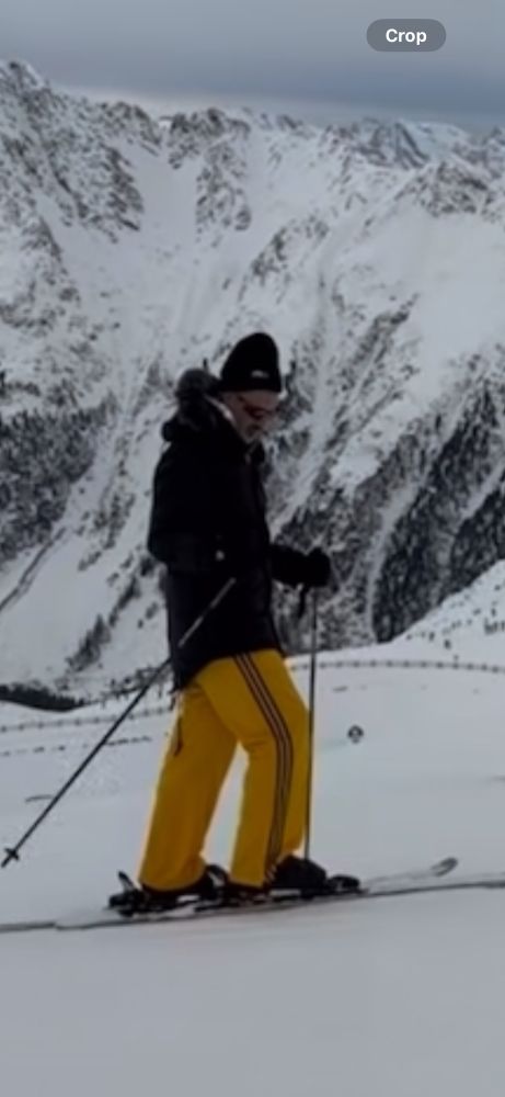 Taika about to go skiing, swinging a ski pole back and forth wearing yellow ski pants with a black stripe, black parka, hat and sunglasses 