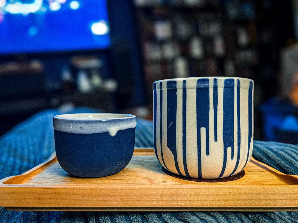 two porcelain mugs sitting side-by-side on a wooden serving tray. The mug on the left is blue with a little bit of white around the lip. It sized for espresso. The other cup on the right has blue streaks, stripping down the side and the rest is white. Around the lip is a clear glaze. It’s sized for cappuccinos.