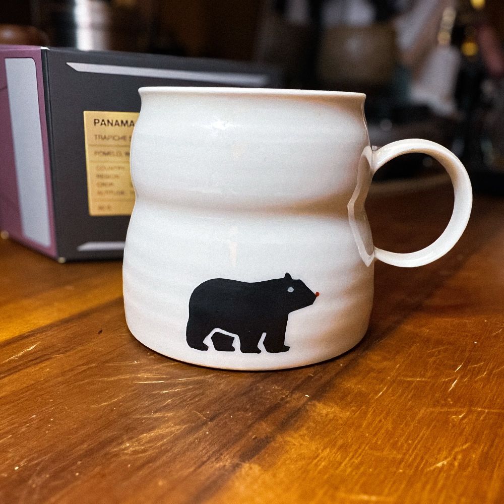 a small white porcelain ceramic mug with handle. on the mug is a black bear with red nose. it’s sitting in a wooden cutting board