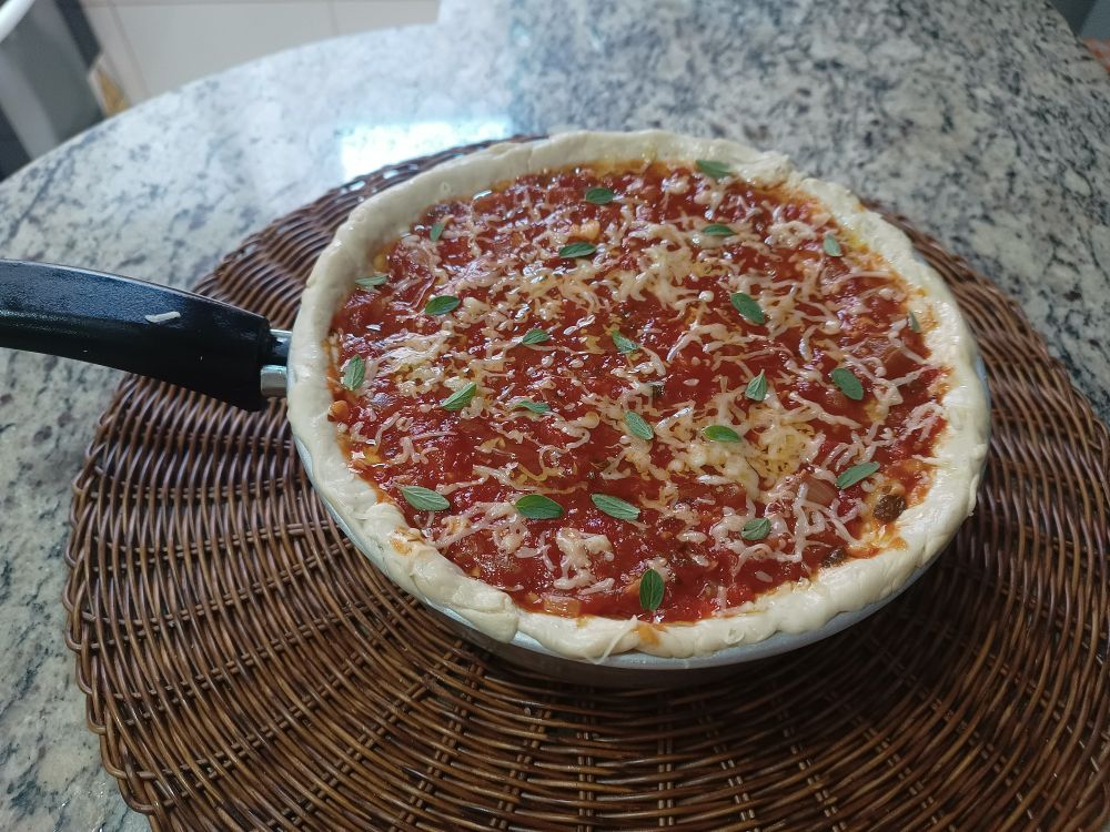 pizza estilo chicago ainda crua cobrindo toda uma frigideira

no topo é possível ver uma borda de massa, molho vermelho com queijo ralado e folhas de manjerona