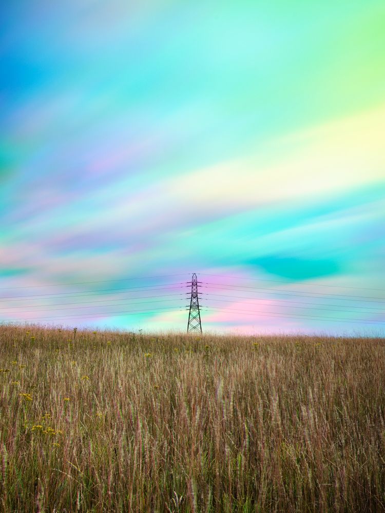 A solitary electricity pylon stands in the middle of a vast, golden-brown field of long grass under a surreal, multicolored sky. The sky is a blend of pastel blues, pinks, greens, and yellows, with soft, streaky clouds creating a dreamlike effect. The pylon, silhouetted against the vibrant backdrop, has multiple horizontal power lines extending from either side, disappearing into the distant horizon. The field fills the bottom two-thirds of the image, with small specks of yellow flowers visible in the foreground.