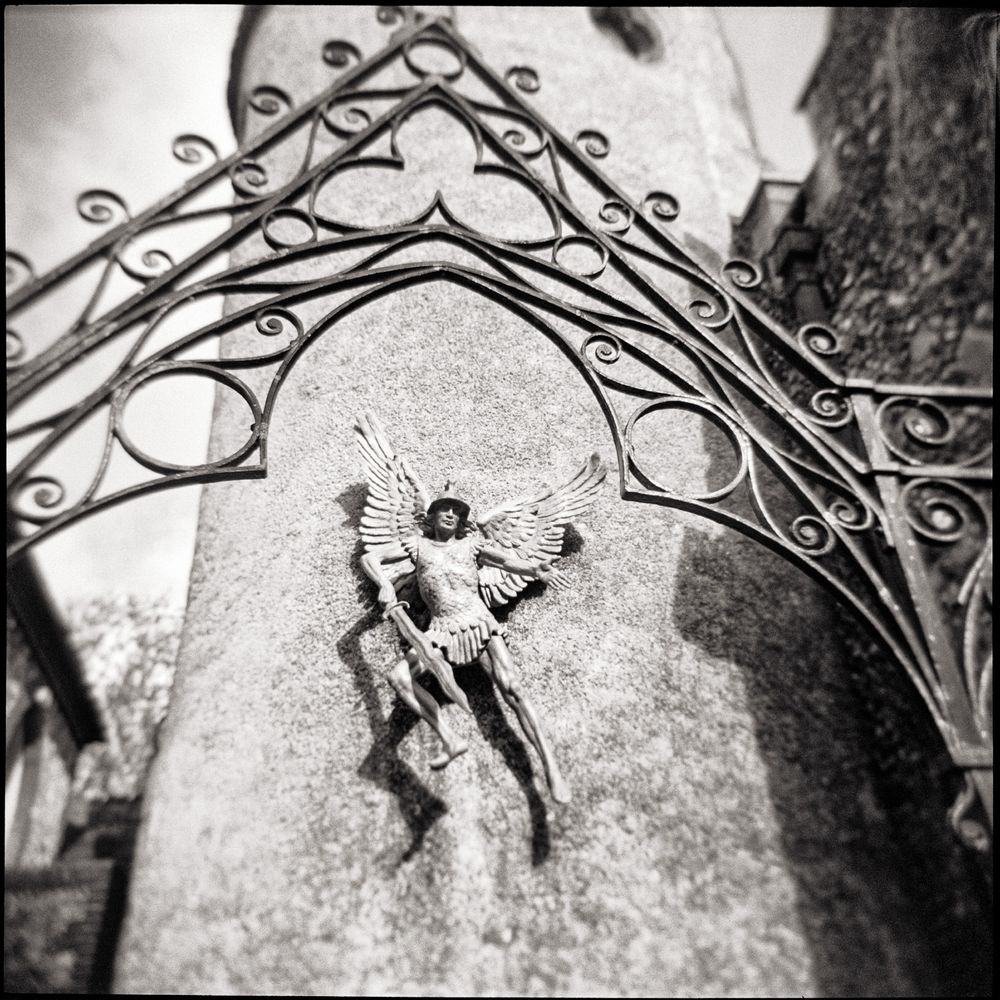 An antique, square-format, black and white photograph captures a small, metal relief sculpture of an angel mounted on a textured stone wall, likely a cemetery gatepost or a church tower. The angel, possibly Saint Michael the Archangel, is depicted in a triumphant pose, slaying a dragon or demon below his feet.

Above the sculpture, a decorative, wrought-iron arch or gatework frames the angel, with intricate scrollwork and pointed Gothic elements that draw the eye toward the central figure. The low-angle perspective emphasizes the height of the post and the dramatic nature of the scene, creating a sense of scale and shadow.