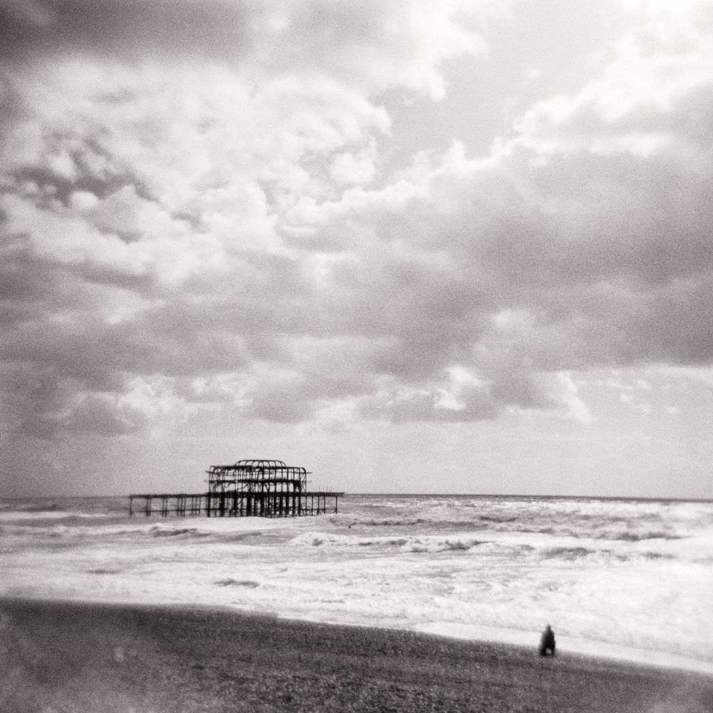 A high-grain, black and white photograph captures the ruins of the West Pier in Brighton, England, silhouetted against a vast, cloudy sky and the churning sea. The pier's skeletal, burnt-out dome and fragmented supports jut out from the water in the middle distance. In the foreground, the shingle beach slopes down, with a single, small, dark figure standing near the water's edge on the lower right, adding a sense of scale and solitude to the dramatic, textured scene. The photograph evokes a somber, atmospheric mood.