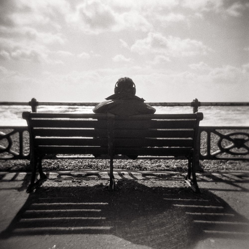 A high-contrast, grainy black and white photograph shows a person seated alone on a wooden bench, facing away from the camera and looking out towards the sea. The person is wearing headphones and a jacket. The bench casts a strong, long shadow towards the camera, emphasizing the bright sun and dramatic sky with scattered clouds. A metal railing separates the person from the water, which appears slightly blurry. The overall feeling is one of solitude and contemplation.