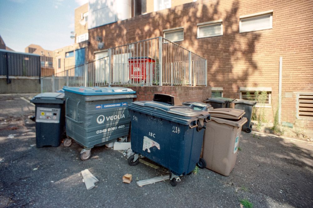 A slightly low-angle photograph captures a cluster of large commercial waste bins in an asphalt service area next to a brick building. The most prominent bin is a large blue Veolia bin marked "CARDBOARD ONLY," standing next to a slightly smaller dark blue IBC bin and a brown bin. A black bin stands on the far left. The bins are on wheels, and some scattered trash is on the ground. A metal railing surrounds a raised landing leading to the reddish-brown brick building.